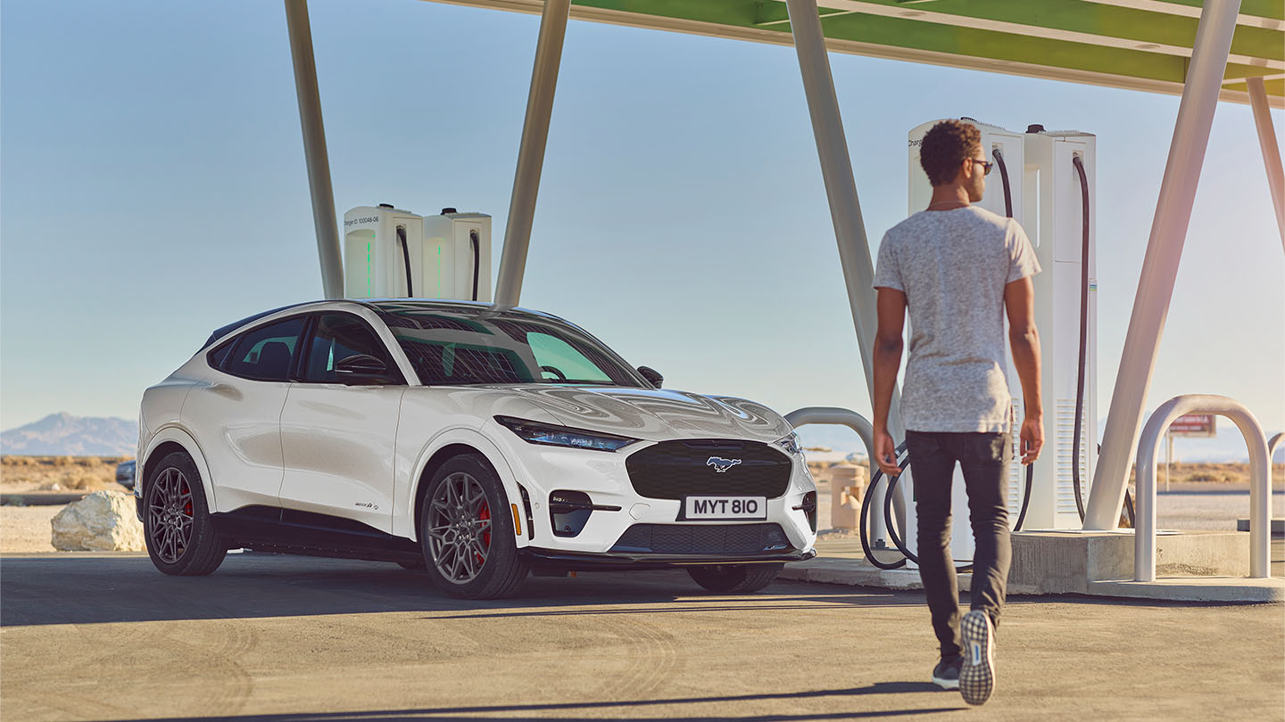 Ford Mustang Mach-E branco estacionado em estação de carregamento exterior. Homem de t-shirt branca caminha para o carro.