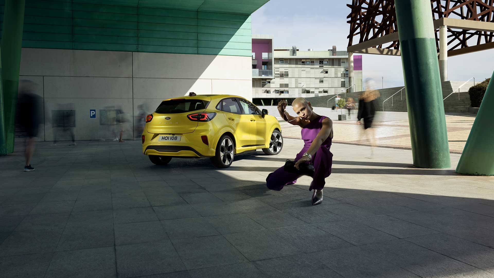 Vista traseira de um Ford Puma Gen-E® elétrico amarelo estacionado debaixo de uma ponte, ao lado de uma pessoa vestida de roxo.