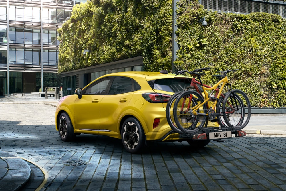 Ford Puma Gen-E amarelo estacionado em ambiente urbano com duas bicicletas montadas num suporte traseiro.