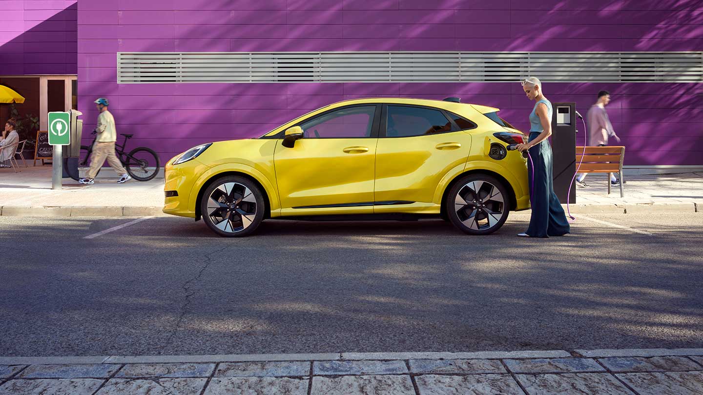 Vista lateral de um Ford Puma Gen-E® elétrico amarelo ligado a uma estação de carregamento pública num lugar de estacionamento.