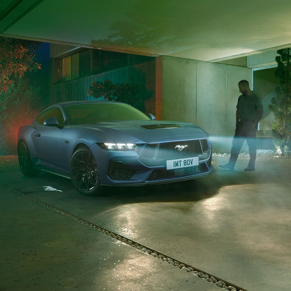 Homem aproximando-se de um Ford Mustang estacionado com os faróis acesos, mostrando a iluminação de boas-vindas ao detetar aproximação